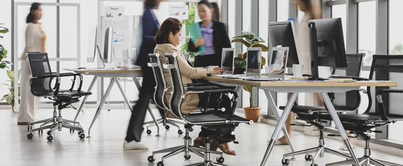 Selective focus on one business woman sitting and working on laptops on conference table with other blurry business women walking in rush in office with multiple glass windows