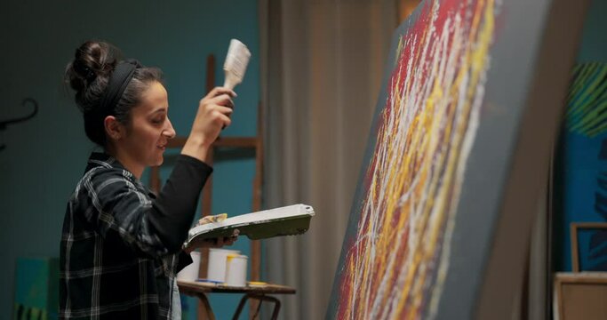 Artist devotes energy to new work of art stands in front of canvas in studio looks at painting raises hand with brush stained with white paint strong quick movements color splashes in front of her