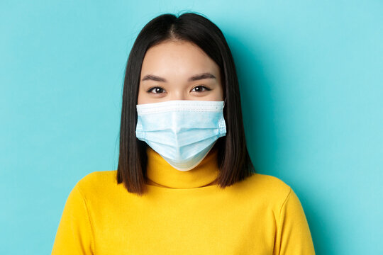 Covid-19, Social Distancing And Pandemic Concept. Close Up Of Young Asian Woman With Short Dark Hair, Wearing Medical Mask And Smiling With Eyes, Looking Hopeful At Camera