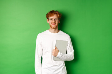 Handsome young man with red hair, wearing glasses and long-sleeve t-shirt, holding laptop on green background