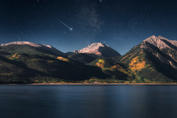 Nighttime scene along the shoreline of a lake with mountains in the background and a shooting star...