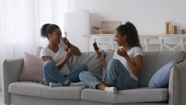 Two Cheerful Black Sisters Eating Ice-Cream And Laughing On Couch At Home
