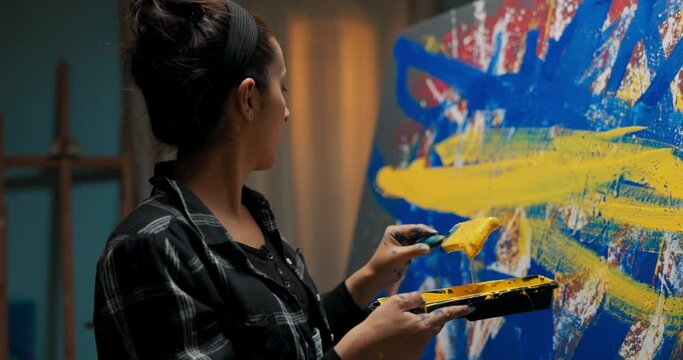 Woman stands at canvas with abstract modern oil painting, girl waits for paint to drip from brush into tray precise strokes of yellow color thick stripes on blue lines