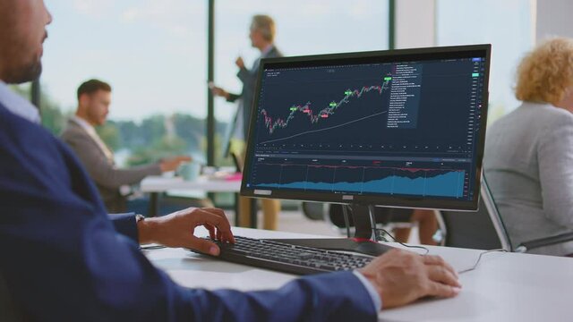 Smiling man working on computer, looking at updated data from stock market. Buy or sell? Modern open space office interior.