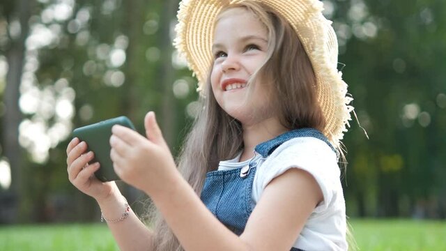 Happy smiling child girl looking in mobile phone outdoors in summer.