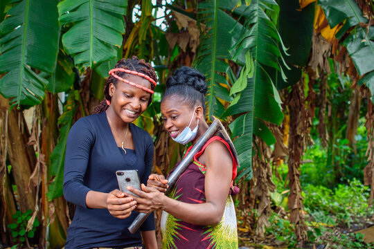 A Female African Farmer With Nose Mask And A Farming Hoe On Her Shoulder Happily Looks Into A Smart Phone With A Female Friend In A Banana Farm Or Plantation