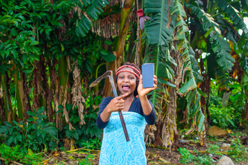 A happy female african farmer with local beads on her head, farming hoe on her shoulder, joyfully...