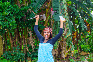 A happy female african farmer with local beads on her head, farming hoe on her shoulder, joyfully raising a farming hoe and smart phone up in a farm