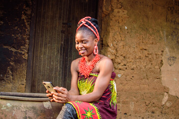 A happy African lady or woman with beads on her head, sitting beside big water calabash, excitedly...