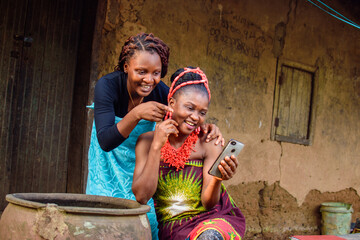 Two female Africans happily looking into the smart phone outside a village mud house with a big...