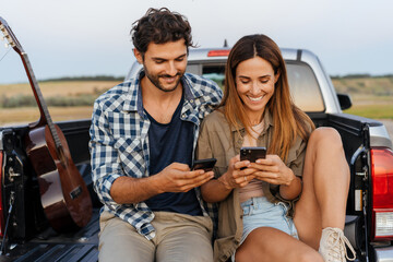 White couple smiling and using cellphones while sitting in car trunk