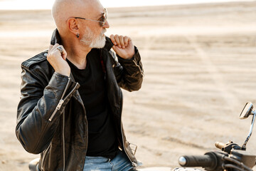 Bold senior man wearing leather jacket posing on motorcycle outdoors