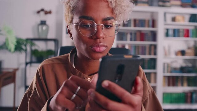 Attractive African American woman sits indoors holding in hands smart phone dial number on touchscreen. Young bicultural female surfing internet, websurfing website, choose buying goods online concept