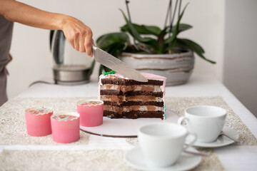 Piece chocolate cake with many layers of cream decorate with pink mastic. Cut piece. Hand cuts cake by big knife. Two white cups of tea on table background. Cake on holiday.