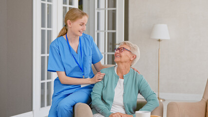 Young nurse in uniform listens to senior patient while sitting on armrest of comfortable armchair - 459842251