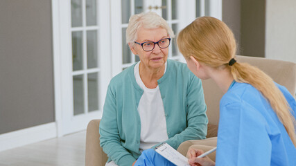 Young blonde doctor interviews short haired senior lady patient making notes in pad at appointment