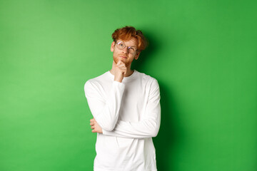 Thoughtful redhead man in glasses making choice, looking up and thinking, standing over green background