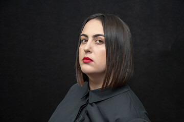 Studio portrait of a plump, round-faced woman 30-35 years old with black hair on a black background, with brightly painted red lips, dark tone. Perhaps she is a director or a television announcer, 