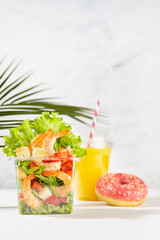 Healthy fresh summer tropical breakfast take away  - set of orange juice, salad with shrimps, red pepper, lettuce, croutons, pink donut in plastic pack in white kitchen interior, palm leaf, vertical.