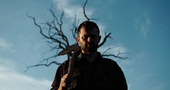 One handsome strong stylish male logger of young man with long lush black beard and moustache in shirt holding wooden axe walking in forest outdoor on natural background, square picture.