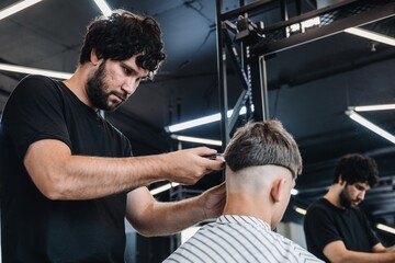 Stylish bearded brunette barber makes a fashionable haircut to a young guy. People at work. Small...