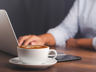 Close-up of a white coffee cup on a wooden table. Women working on a laptop and drinking coffee in the morning relax mood. Beverage, lifestyle, and relaxation Concept