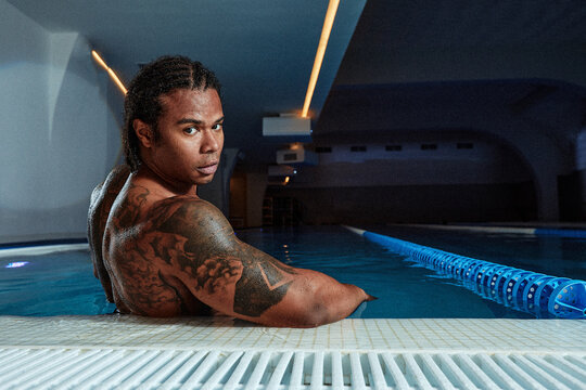 Young hot man resting at swimpool. Back view of guy sitting at water alon. Enjoy his time lonely in resort spa place. Strong powerful man.