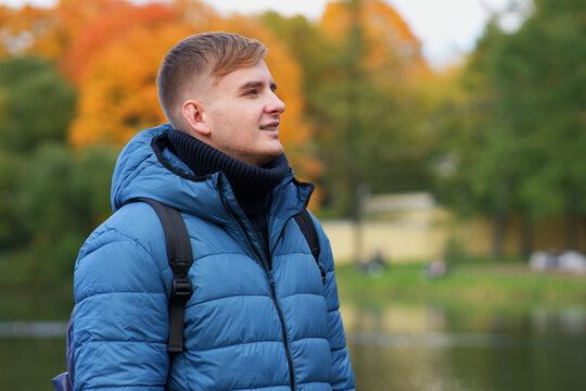 Portrait of handsome positive happy guy, young man in sweater, down jacket with backpack standing, walking in beautiful golden autumn park, smiling. Natural background, colourful leaves, trees.