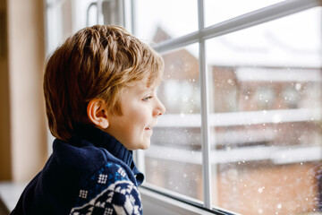Happy adorable kid boy sitting near window and looking outside on snow on Christmas day or morning....