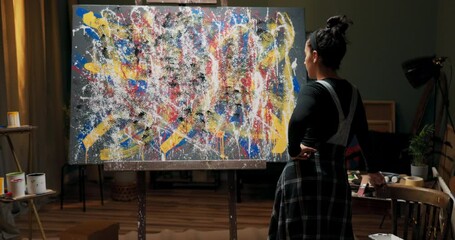 Talented pretty girl is standing in art studio leaning against table with brushes and paints, looking at her work, painting, admiring abstract combination of colors, contemplating improvements