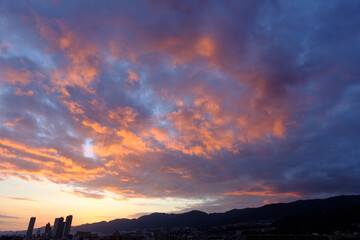 都会の夕暮れ。六甲山に沈む夕陽とオレンジ色に染まる空と雲。高層ビルがシルエットで浮かぶ。