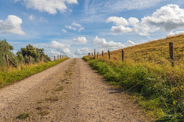 Fototapeta na wymiar Driveway of a dike along the Dutch river Amer near the village of Drimmelen, province of North Brabant. It is a sunny day with a blue sky and cumulus clouds at the beginning of the autumn season.