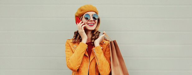 Autumn portrait of happy smiling young woman with shopping bag calling on smartphone wearing an yellow jacket, beret on gray background