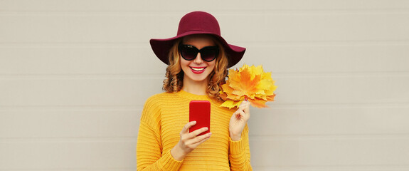 Autumn portrait of happy smiling young woman with smartphone and yellow maple leaves wearing hat on gray background