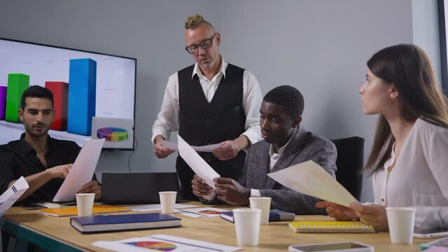 Portrait of serious boss passing paperwork to employee talking. Confident male CEO discussing idea with African American Middle Eastern and Caucasian employees in office meeting room