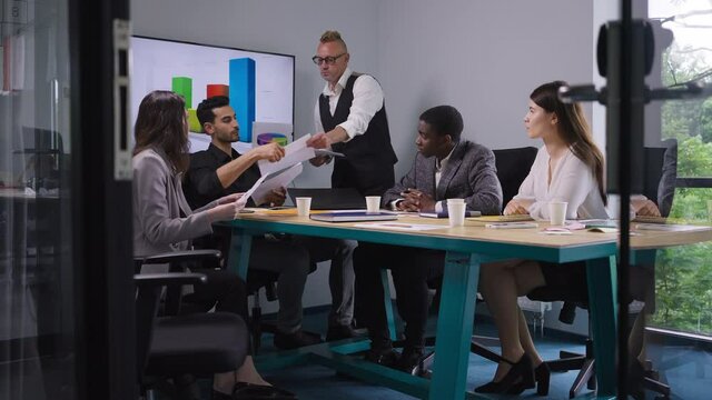 Wide shot senior manager passing documents to multiethnic employees talking explaining strategy idea in office meeting room. Confident professional male CEO brainstorming with colleagues planning
