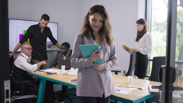 Smiling satisfied Caucasian woman posing with tablet in office with colleagues at background. Portrait of expert manager looking at camera smiling sighing as multiethnic coworkers talking indoors