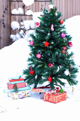 Colorful boxes of gifts on snow and artificial fir tree decorated with Christmas red toys outdoors in yard in snowy background, New Year and Christmas concept