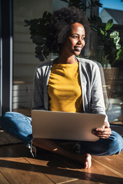 Smiling woman with laptop sitting cross-legged in sunlight at home