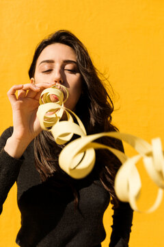 Young woman blowing party blower in front of yellow wall