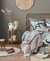 Wooden bed in stylish neutral bedroom interior with design furniture, decoration, carpet, book, dried flowers in vase, bed sheets, blanket, pillows and elegant personal accessories in home decor.