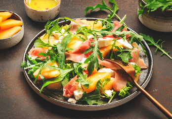 Salad with melon, jamon, goat cheese and arugula on brown background, top view, copy space
