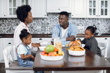 African american parents and their two little daughters sitting on modern kitchen, eating and drinking. Positive family of four enjoying tasty breakfast together at home.