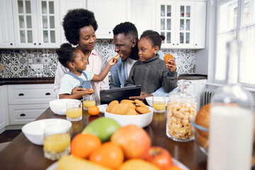 Beautiful african american family of four using digital tablet during breakfast on bright kitchen. Happy young parents watching cartoons with their two daughters while eating at home.