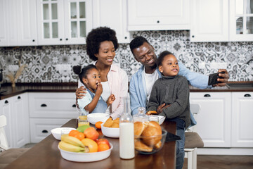 Beautiful african family taking selfie while sitting on kitchen with fresh fruits and cookies. Young parents with two pretty daughters using modern smartphone during breakfast at home.