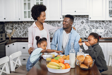 Two cute sisters and their young happy parents eating healthy food on bright kitchen during morning time. African american family chatting and smiling during breakfast at cozy home.