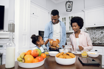 Young african parents and their cute daughter having breakfast with fruits and freshly baked buns. Happy family eating together at home. Enjoyment from morning time and no hurry.