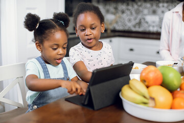 Pretty african american girls watching cartoons on digital tablet while sitting together on bright kitchen. Two sisters using modern gadgets during breakfast at home. Children and technologies concept
