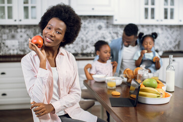 Pleasant african american woman smiling and looking at camera while sitting on kitchen with apple in hands. Husband and two daughters playing on background. Domestic life of happy family.