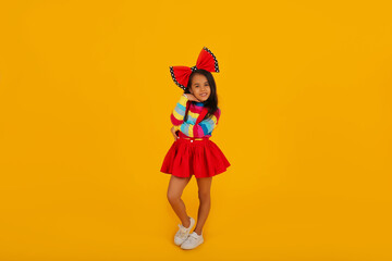 a dark-skinned happy girl in a red skirt in a colored jacket with a red polka dot bow on her head on a yellow background
